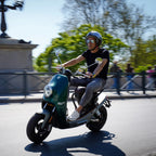 Zöld NITO NES 10 Menet közben, egy középkorú férfi ül a motoron bukósisakban, napszemüvegben a Lánchíd budai hídfőjénél, Budapesten. A háttér elmosódott.
